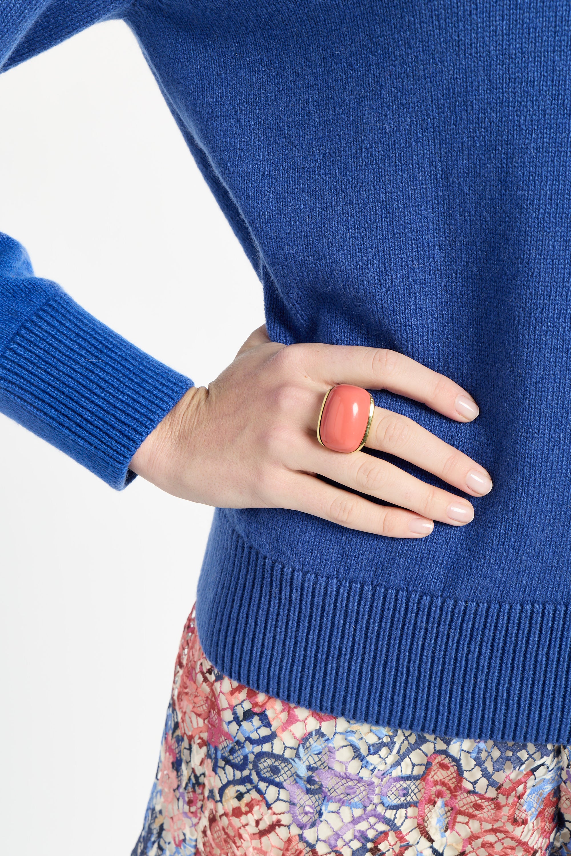 Person wearing a blue sweater with a floral skirt, hand on hip, pink ring on finger.