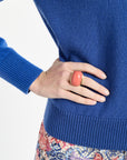 Person wearing a blue sweater with a floral skirt, hand on hip, pink ring on finger.