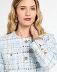 Woman wearing a light blue tweed jacket with gold buttons on a white background
