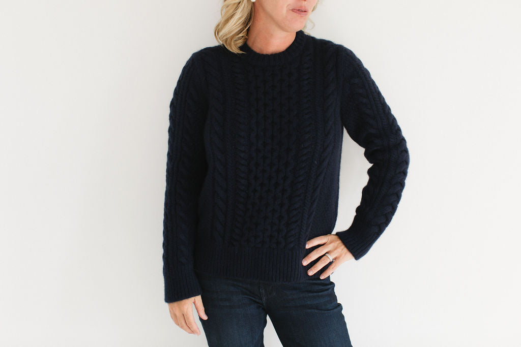 Eleanor Leftwich wearing the cable knit Fisherman Crewneck in navy, paired with dark jeans and pearl earrings against a white background.