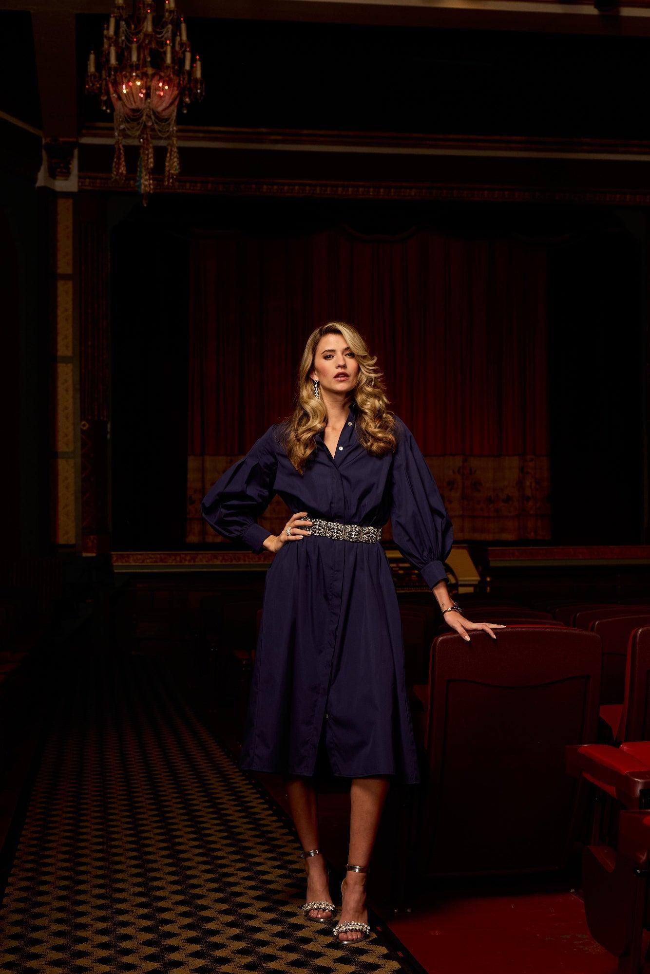 Woman in a dark blue dress standing in a dimly lit room with red curtains and a chandelier.