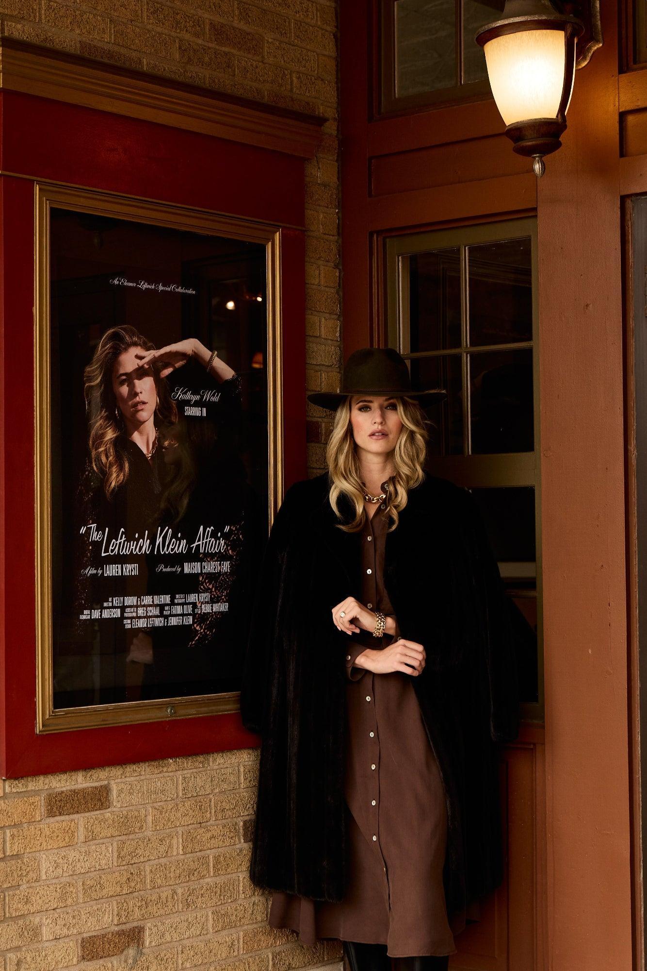 Woman in a coat standing next to a framed poster on a brick wall.