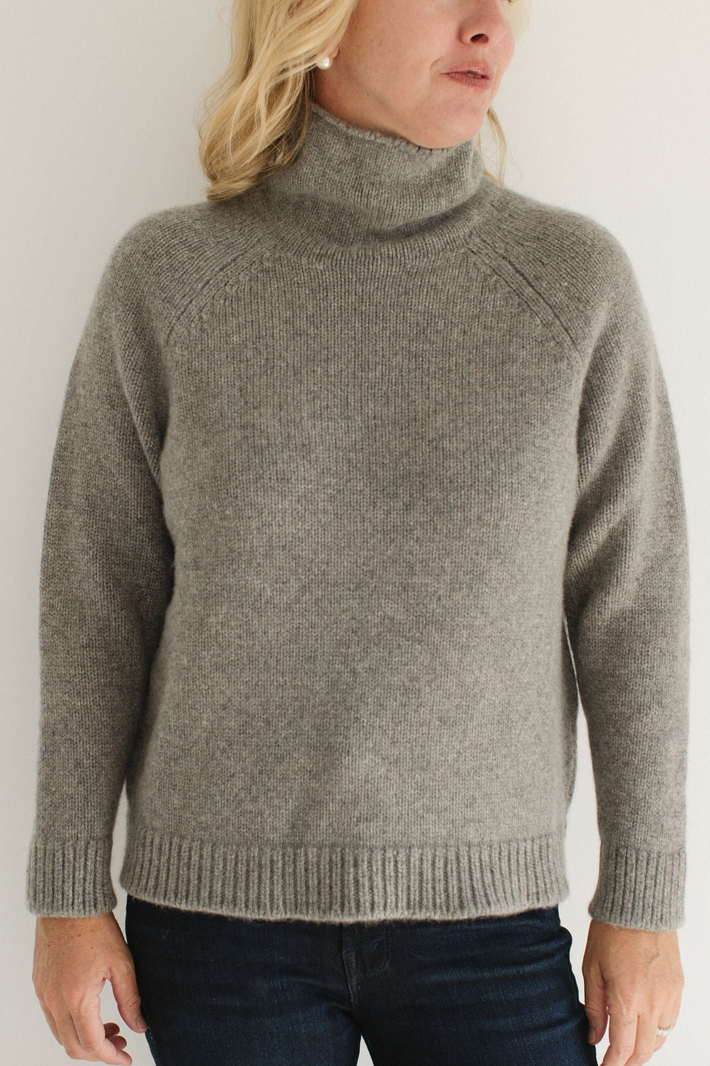 Eleanor Leftwich wearing the Boyfriend Turtleneck sweater in Gray paired with dark jeans and large pearl earrings against a white background.