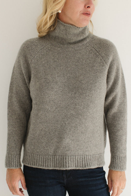 Eleanor Leftwich wearing the Boyfriend Turtleneck sweater in Gray paired with dark jeans and large pearl earrings against a white background.