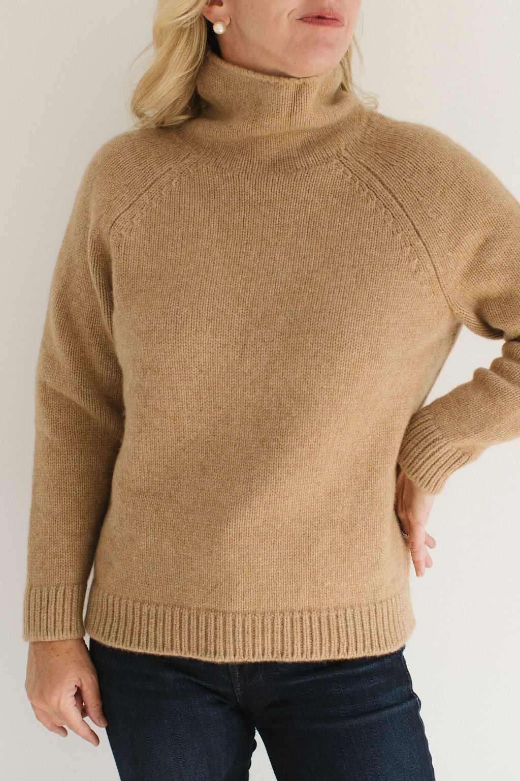 Eleanor Leftwich wearing the Boyfriend Turtleneck sweater in Camel paired with dark jeans and large pearl earrings against a white background.