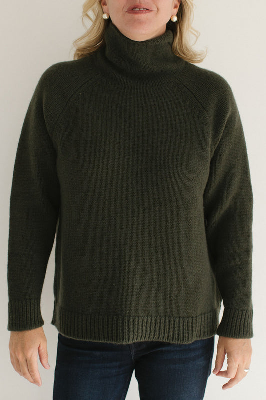 Eleanor Leftwich wearing the Forest Green Boyfriend Turtleneck paired with dark jeans and large pearl earrings against a white background.