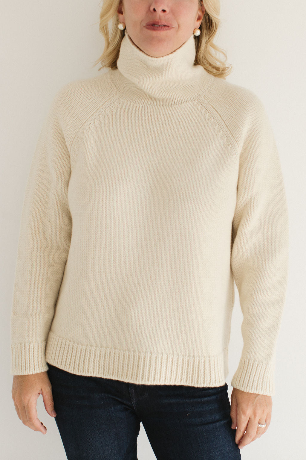 Eleanor Leftwich wearing the Boyfriend Turtleneck sweater in Cream paired with dark jeans and large pearl earrings against a white background.