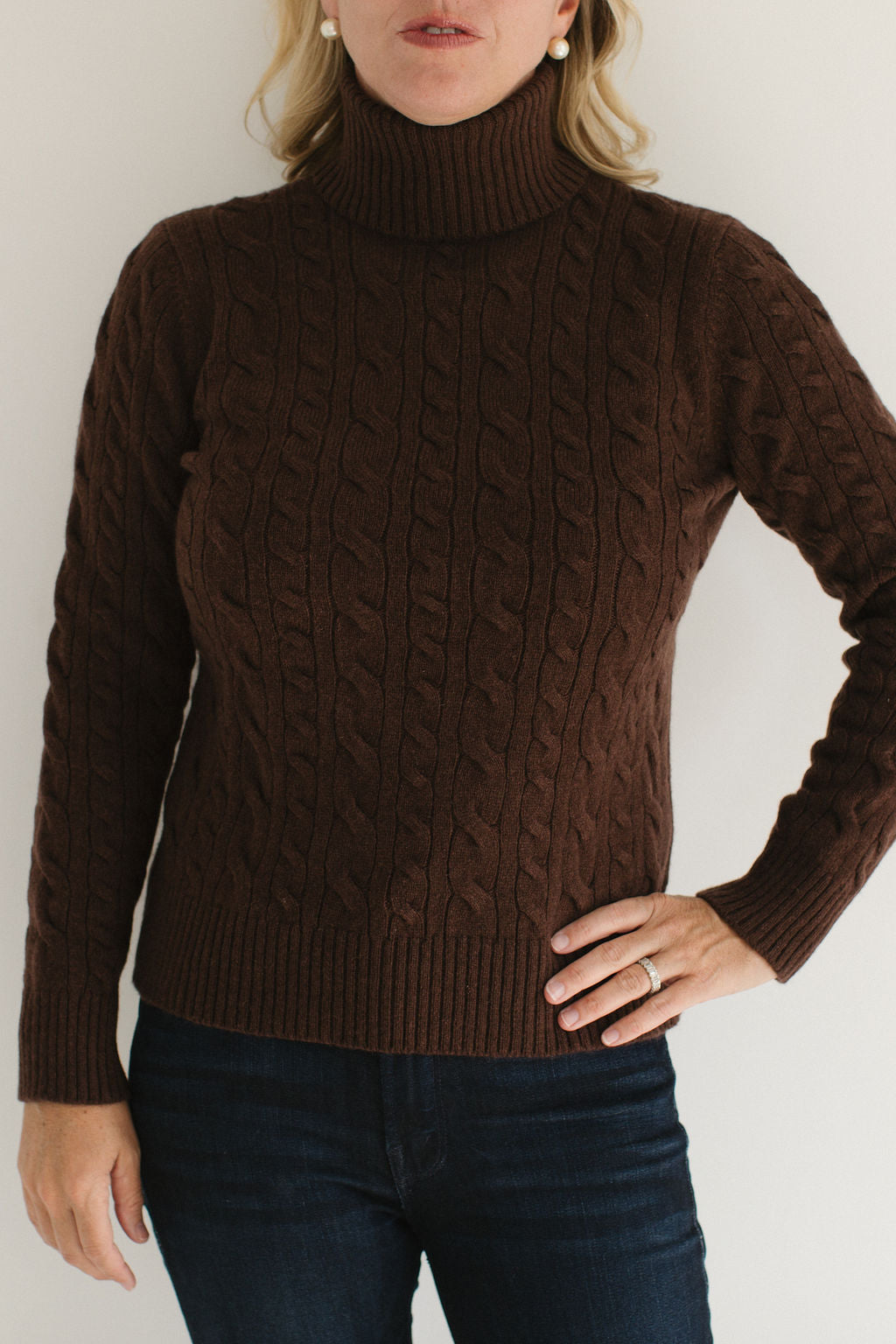 Eleanor Leftwich modeling the Brown Cable-Knit Turtleneck sweater paired with dark denim and gold earrings against an off-white background.