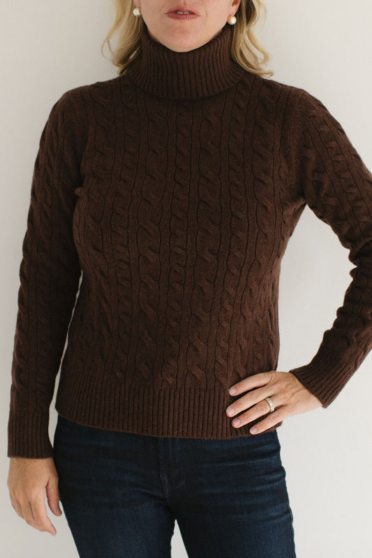 Eleanor Leftwich modeling the Brown Cable-Knit Turtleneck sweater paired with dark denim and gold earrings against an off-white background.