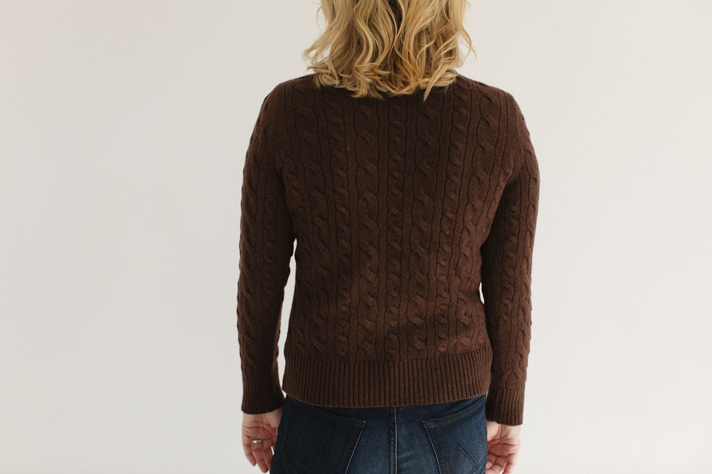 Back view of Eleanor Leftwich in the Brown Cable-Knit Turtleneck sweater paired with dark denim against an off-white background.