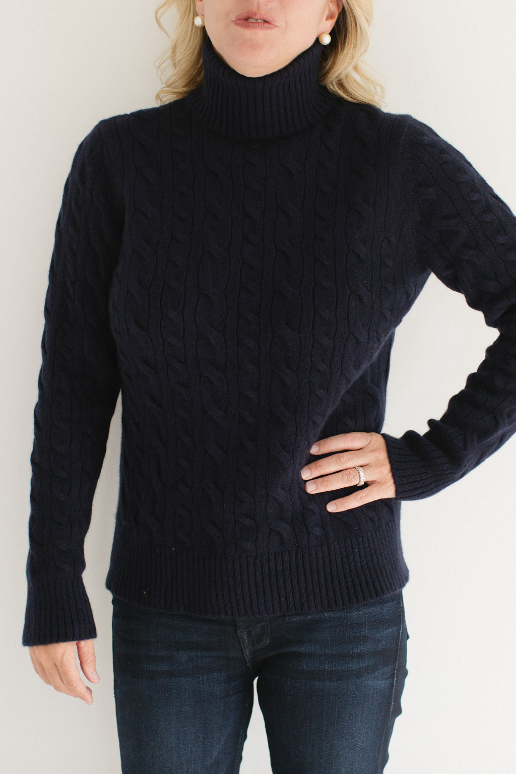 Eleanor Leftwich modeling the Navy Cable-Knit Turtleneck sweater paired with dark denim and gold earrings against a white background.