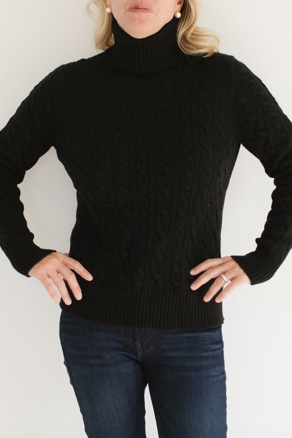 Eleanor Leftwich modeling the Black Cable-Knit Turtleneck sweater paired with dark denim and gold earrings against a white background.