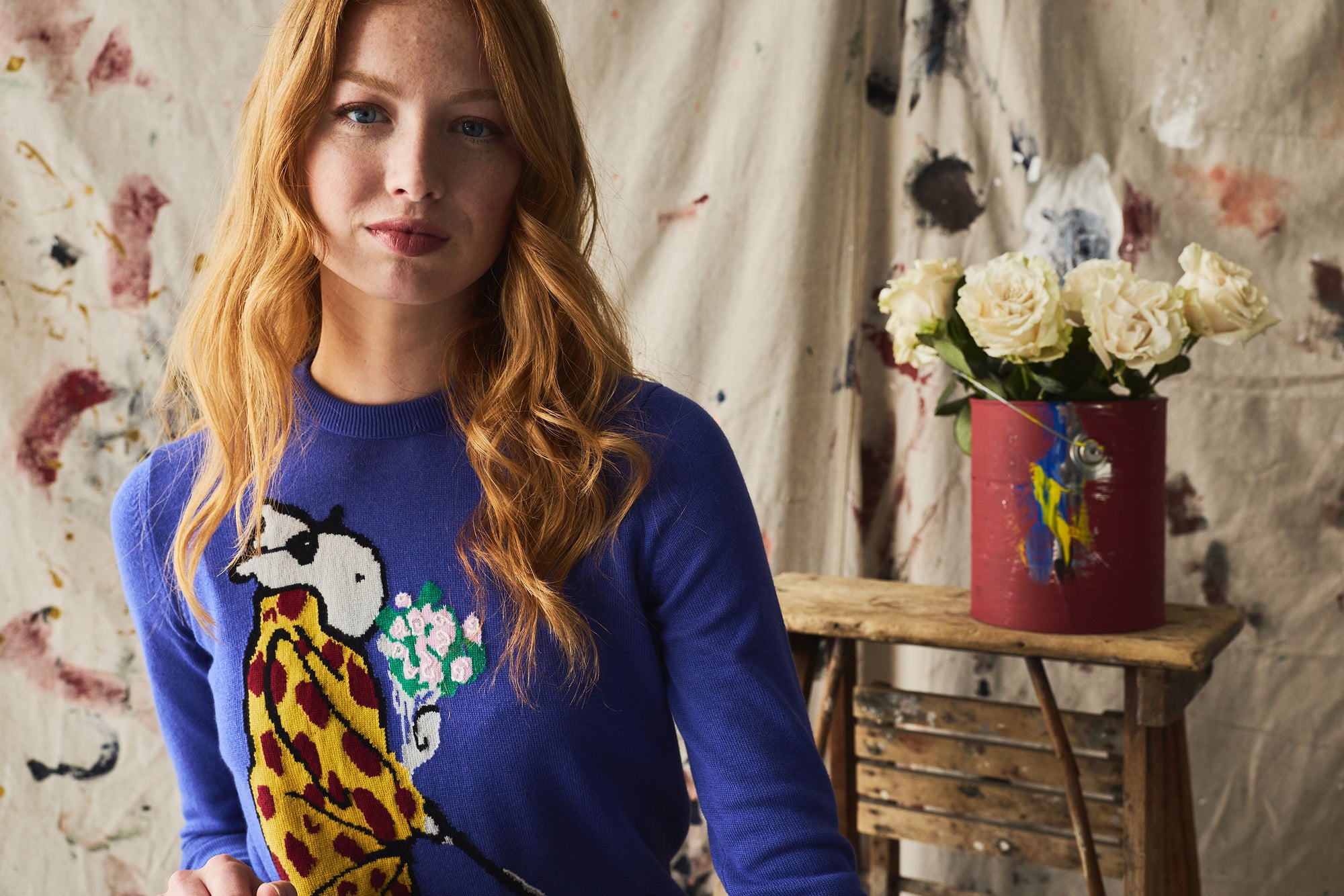 Woman wearing a blue sweater with a colorful design, standing in front of a wall with floral patterns and a small table with flowers.