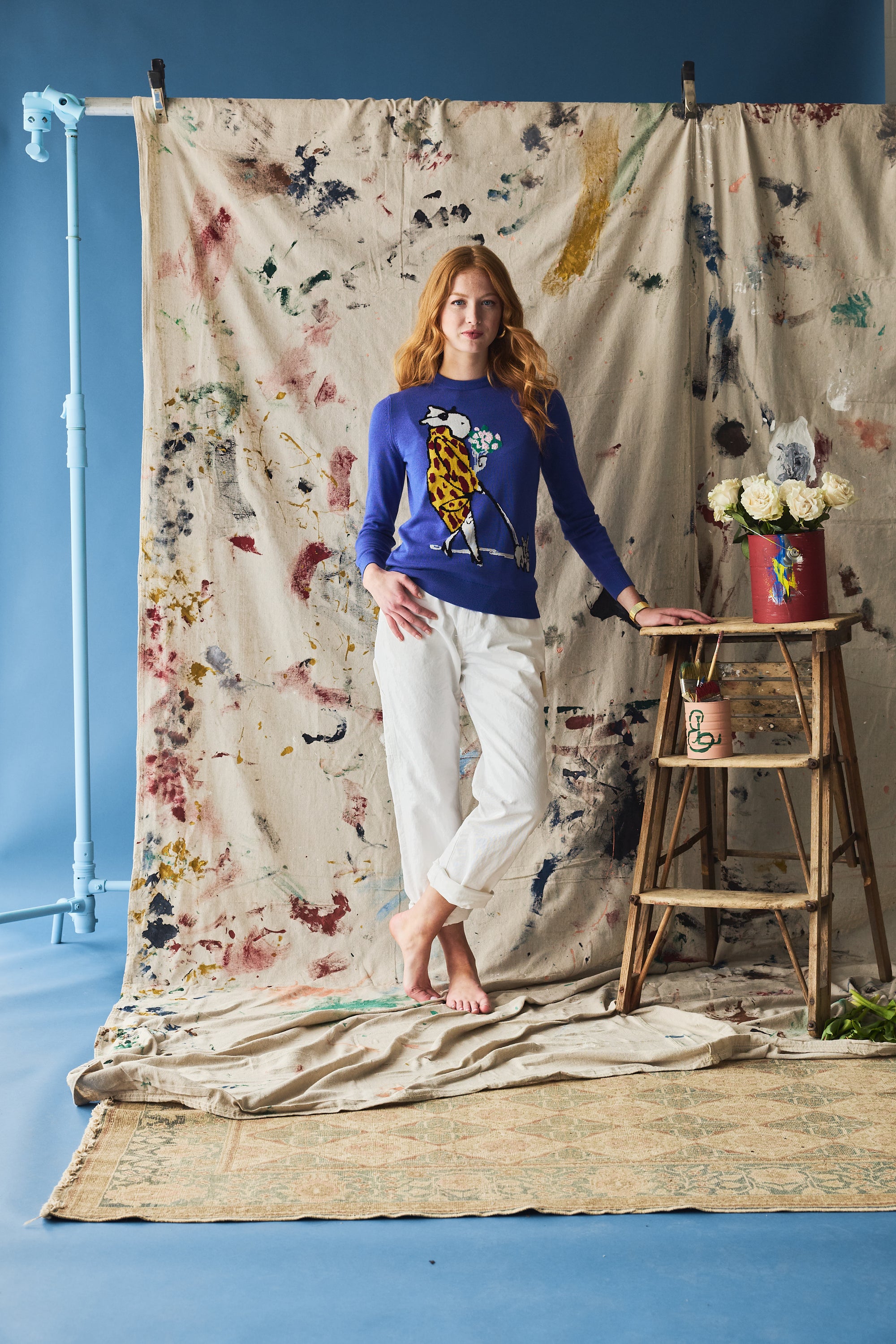 Woman in a blue sweater and white pants standing in front of a colorful fabric backdrop with a ladder and flowers.