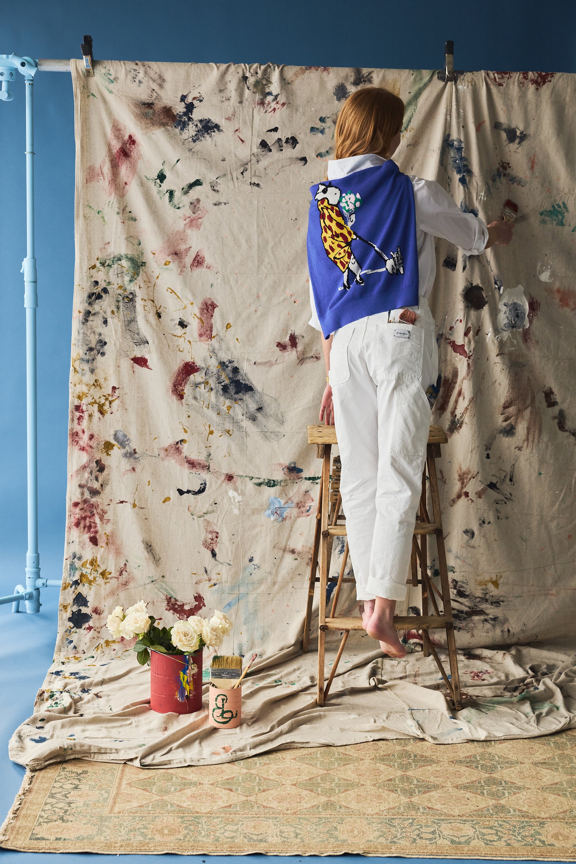 Person standing on a ladder against a large painted backdrop with a blue sweater with a character design on her back