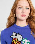 Woman wearing a blue sweater with a colorful design of a hippo and flowers on a white backdrop