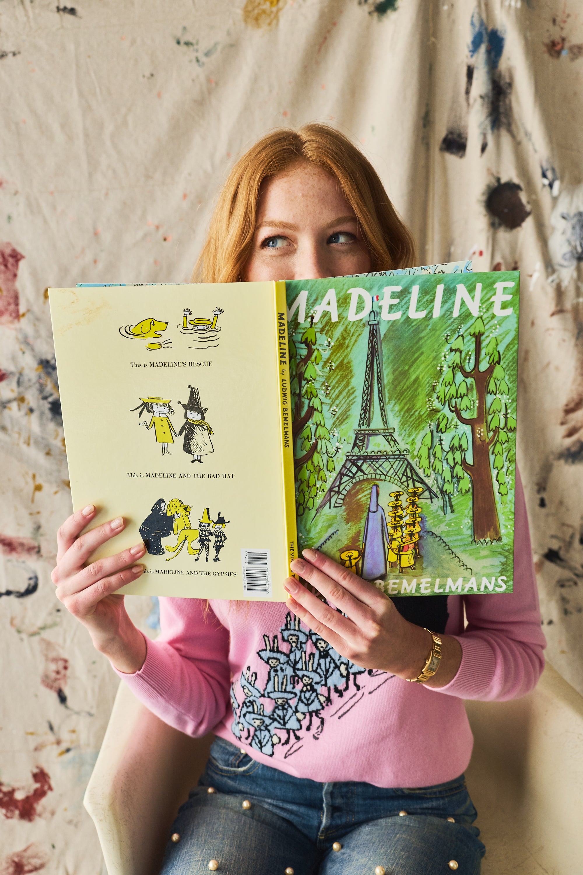 Person holding a book titled 'Madeline' wearing a pink sweater with a paint splotch backdrop