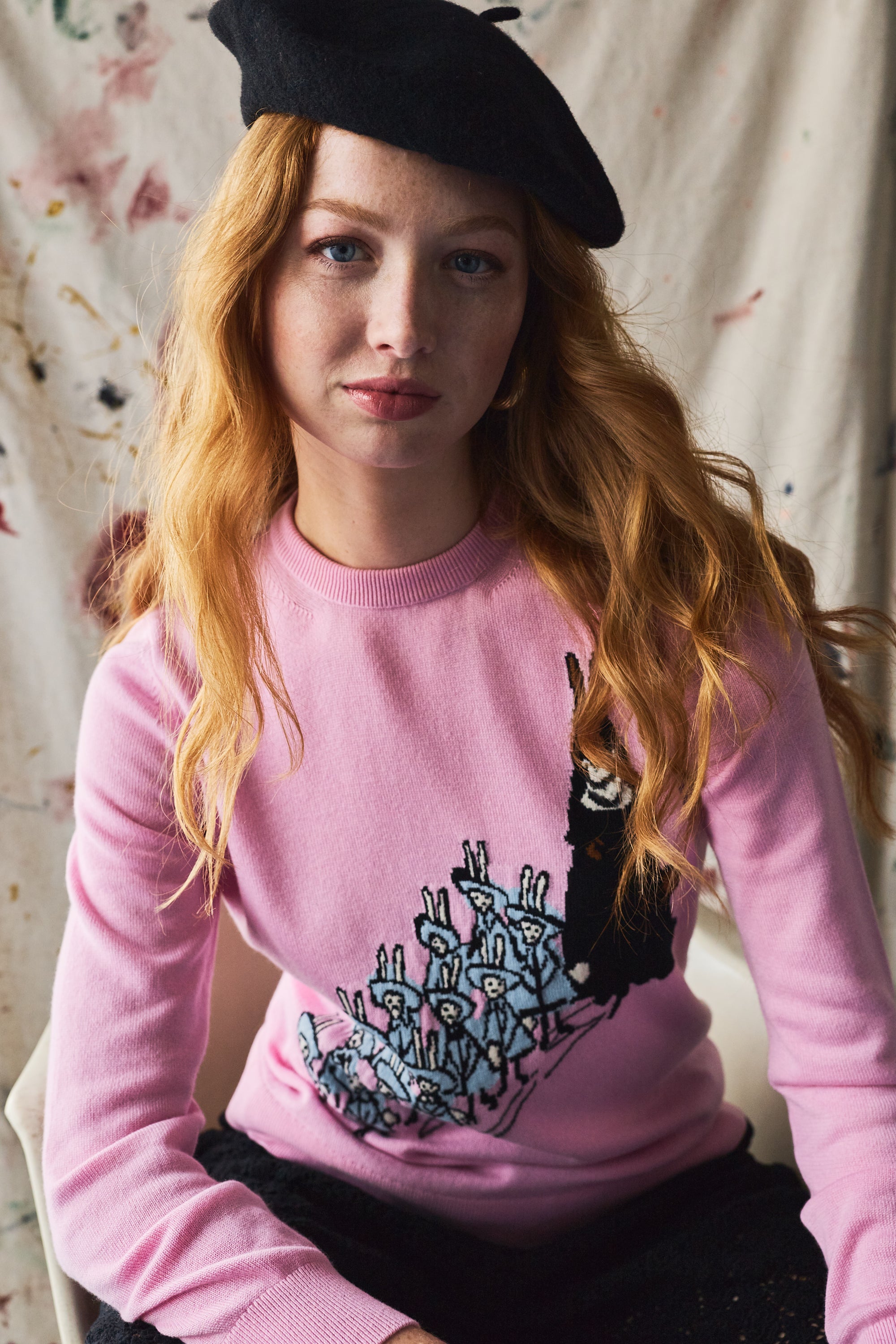Person wearing a pink sweater with a knitted design, sitting against a painted background.