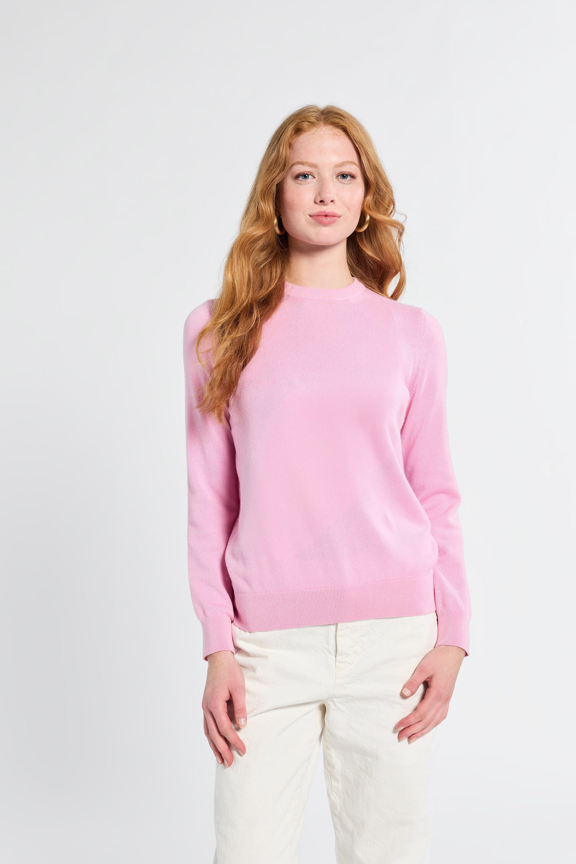 Woman wearing a pink sweater and white pants against a light gray background