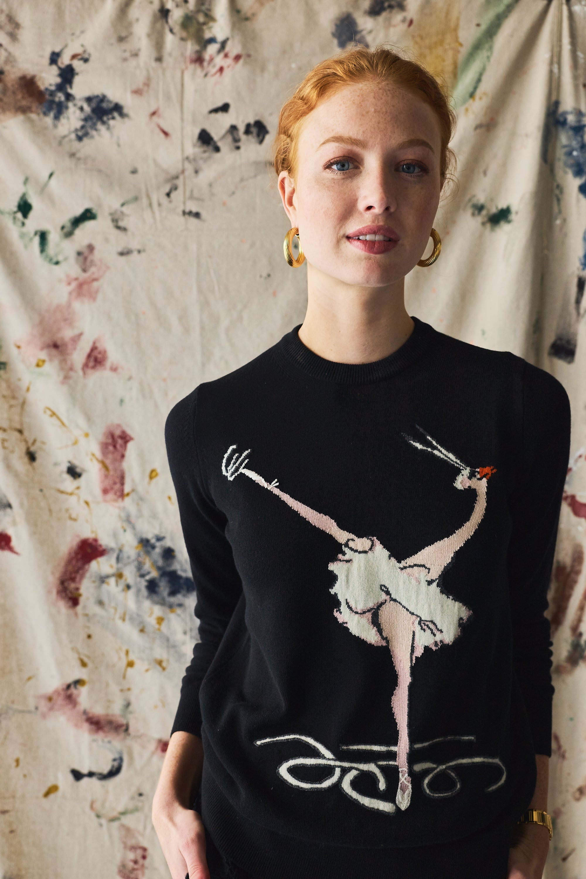 Person wearing a black sweater with a graphic design of a dancer and a bird on a paint patterned wall.