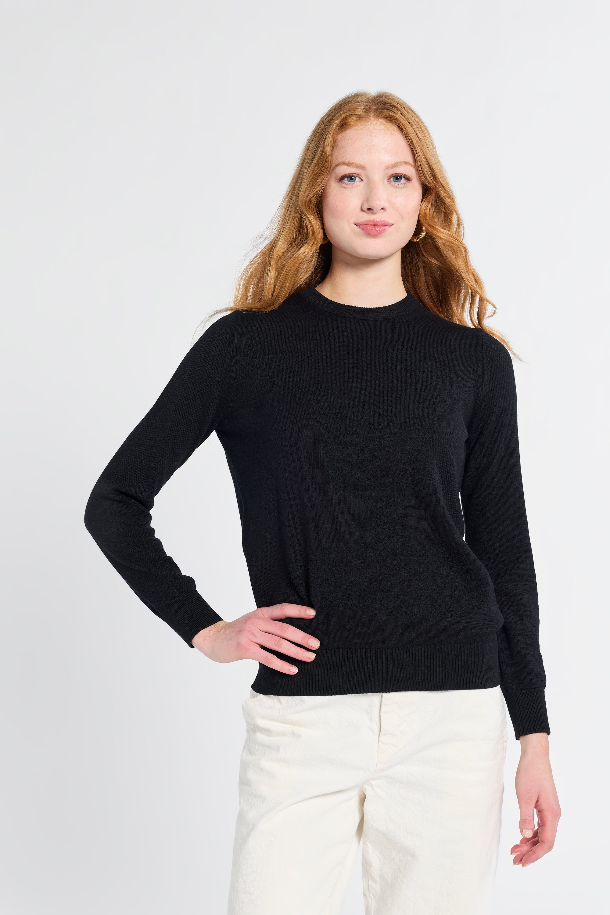 Woman modeling a black sweater wearing white pants on a plain backdrop