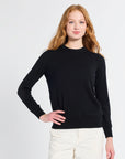 Woman modeling a black sweater wearing white pants on a plain backdrop