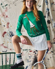 Person wearing a green sweater with a giraffe design, white skirt, and holding a tennis racket against a colorful abstract background.