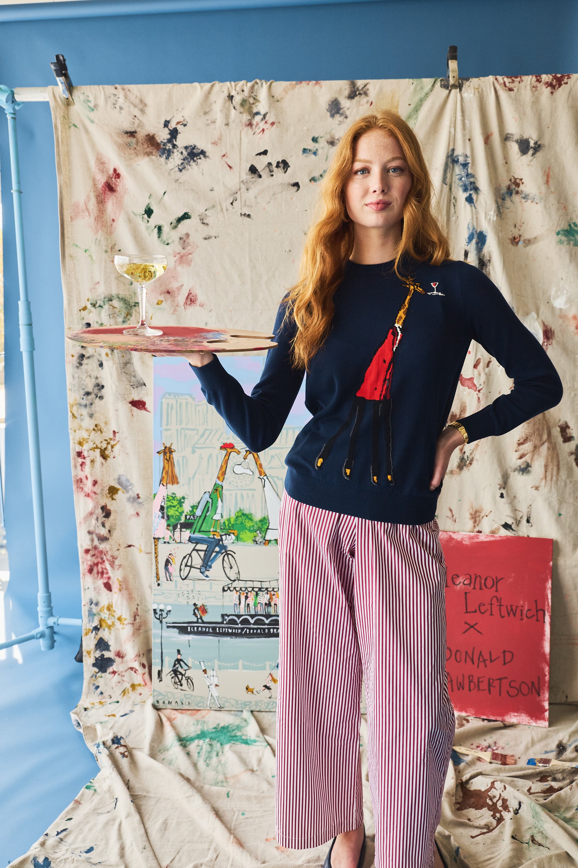 Woman holding a tray with a drink, wearing a navy sweater with a giraffe on it, standing in front of a colorful backdrop