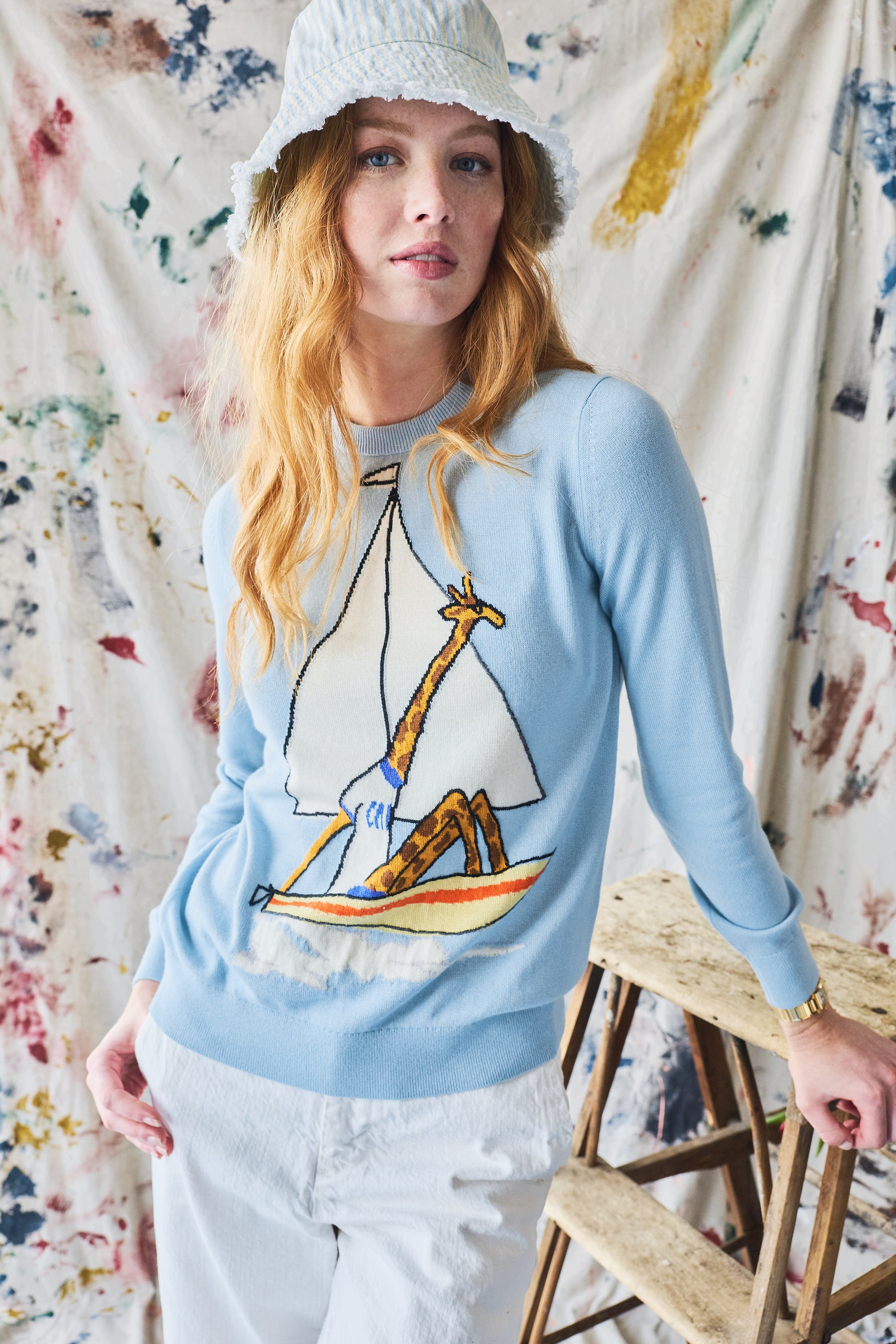Woman wearing a light blue sweater with a sailboat and giraffe design, standing against a paint splotched curtain.