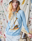 Woman wearing a light blue sweater with a sailboat and giraffe design, standing against a paint splotched curtain.