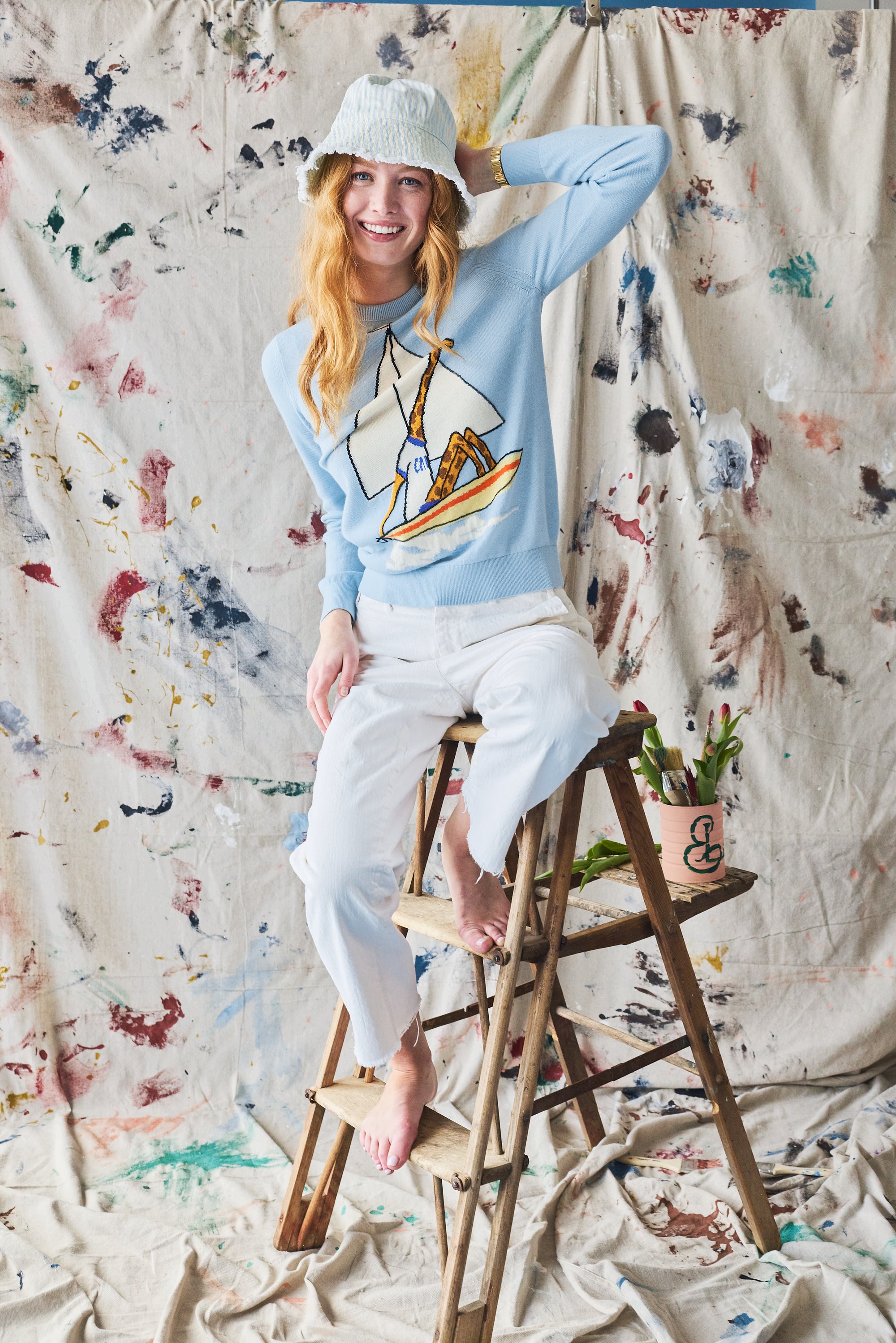 Person sitting on a stool wearing a light blue sweater with sailboat design and white pants, against a textured wall.