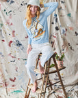 Person sitting on a stool wearing a light blue sweater with sailboat design and white pants, against a textured wall.