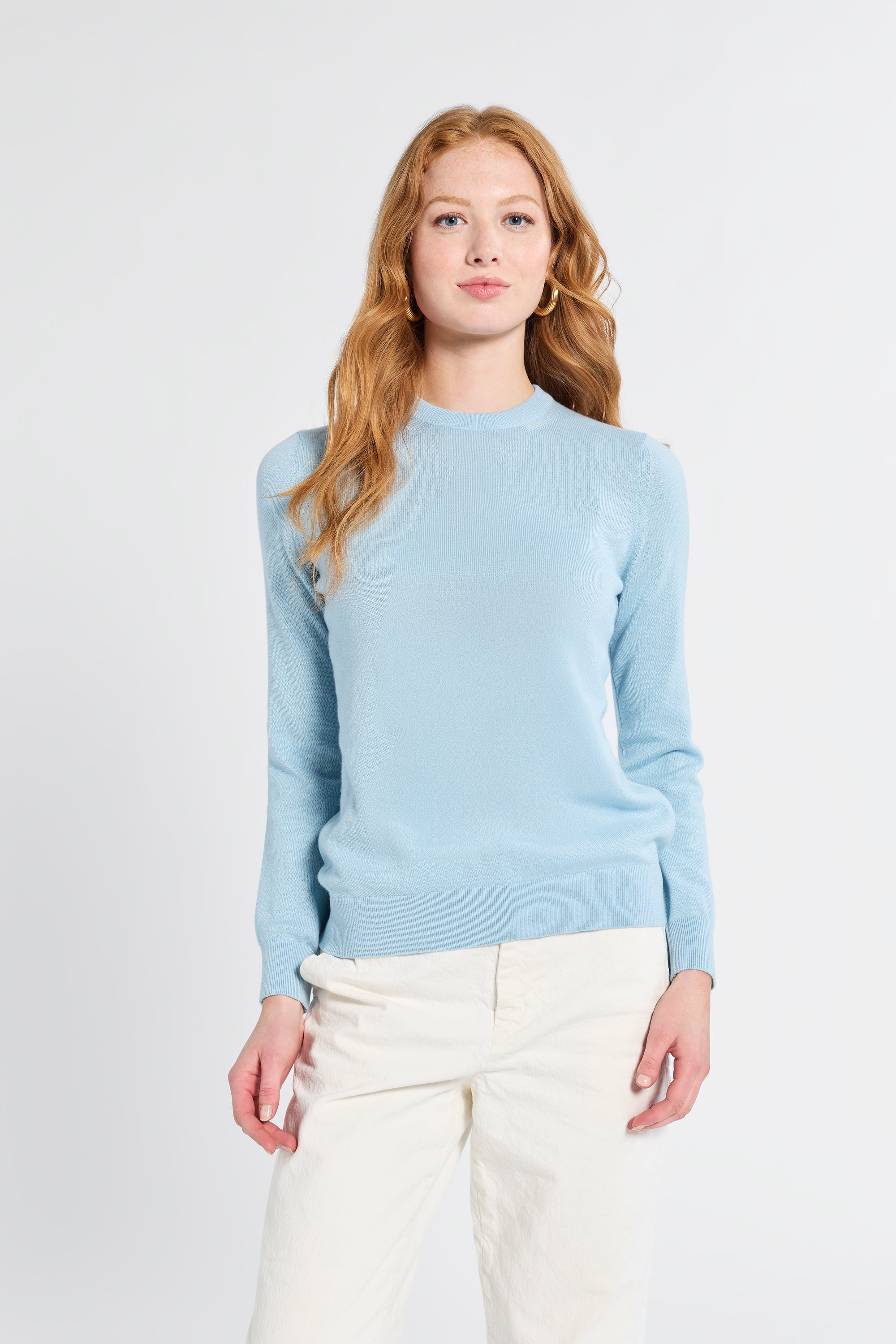 Woman wearing a light blue cashmere sweater and white pants on a plain background