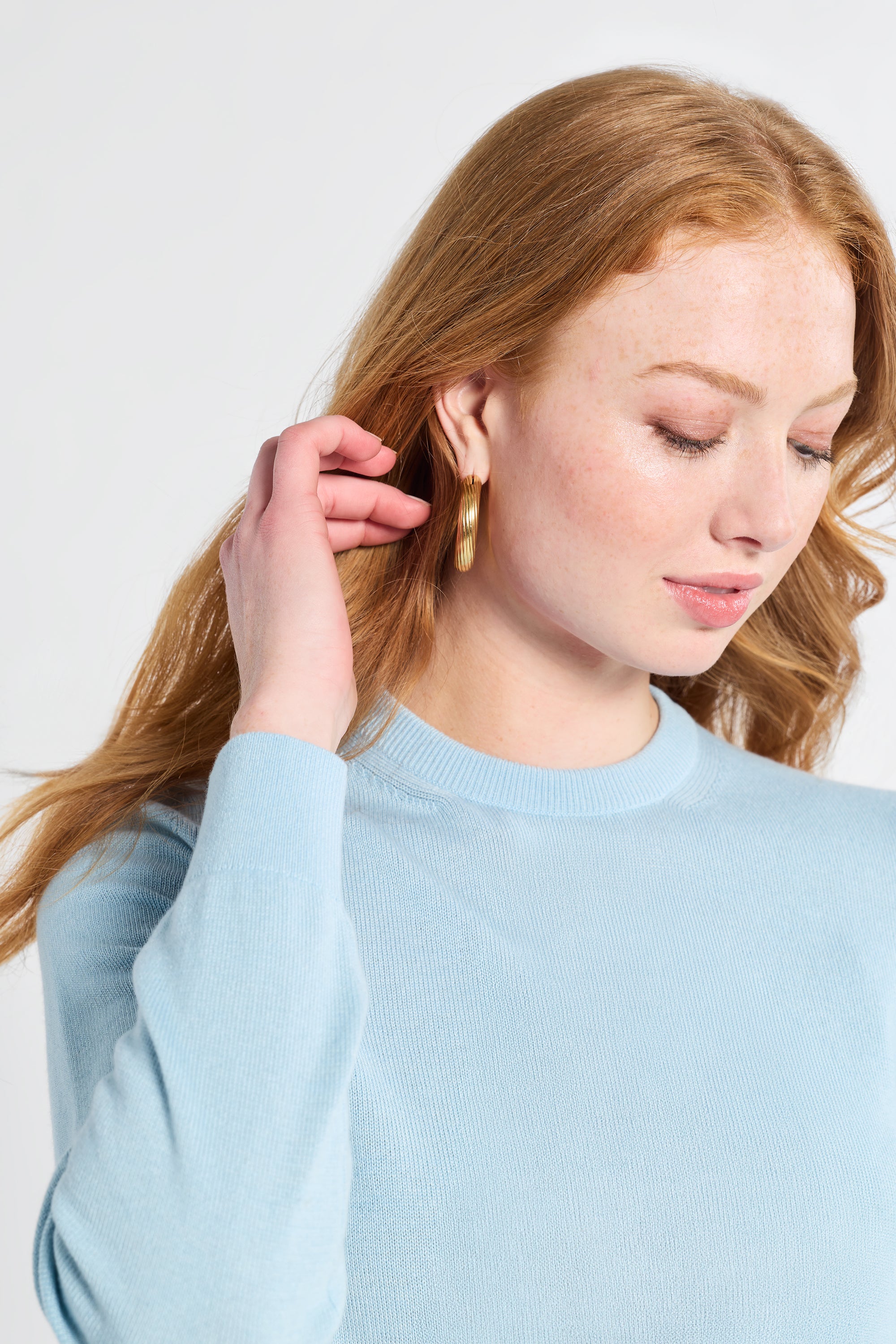 Woman wearing gold hoop earrings and a long sleeve blue silk sweater against a plain background
