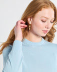 Woman wearing gold hoop earrings and a long sleeve blue silk sweater against a plain background