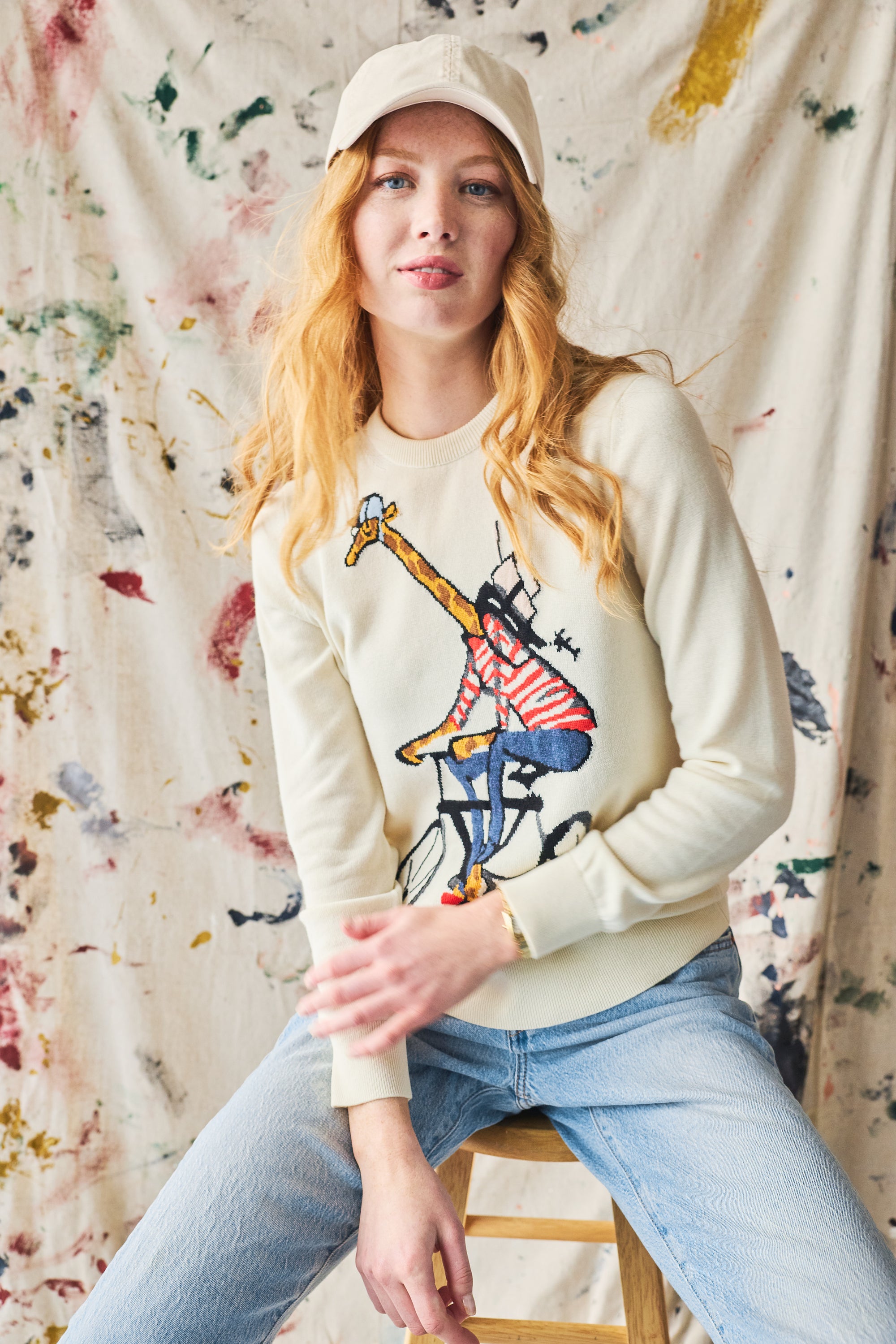 Person wearing a cream sweatshirt with a giraffe design, sitting on a stool against a painted background.
