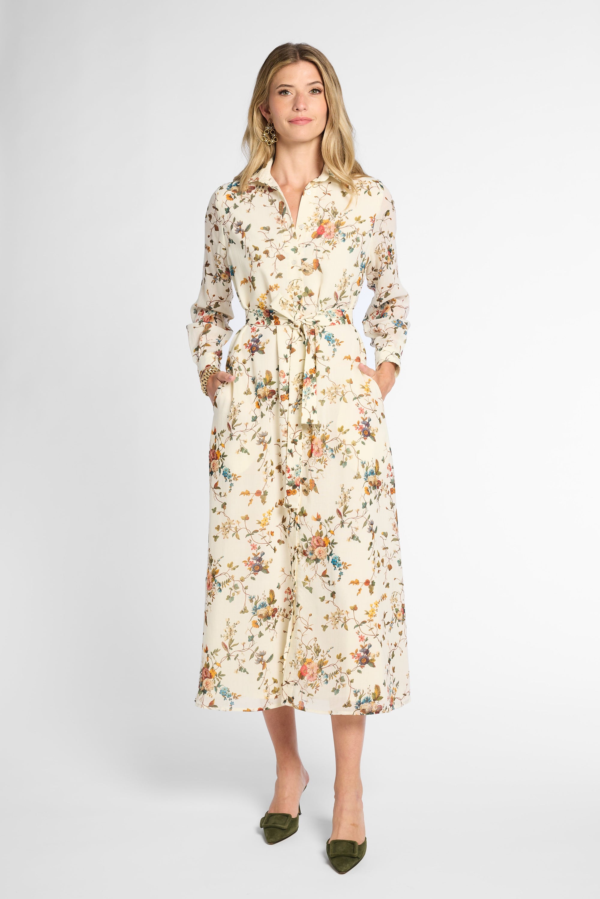 A woman modeling a long-sleeved midi shirt dress with a floral print on a cream background, paired with olive green pointed-toe flats, against a light gray studio background.