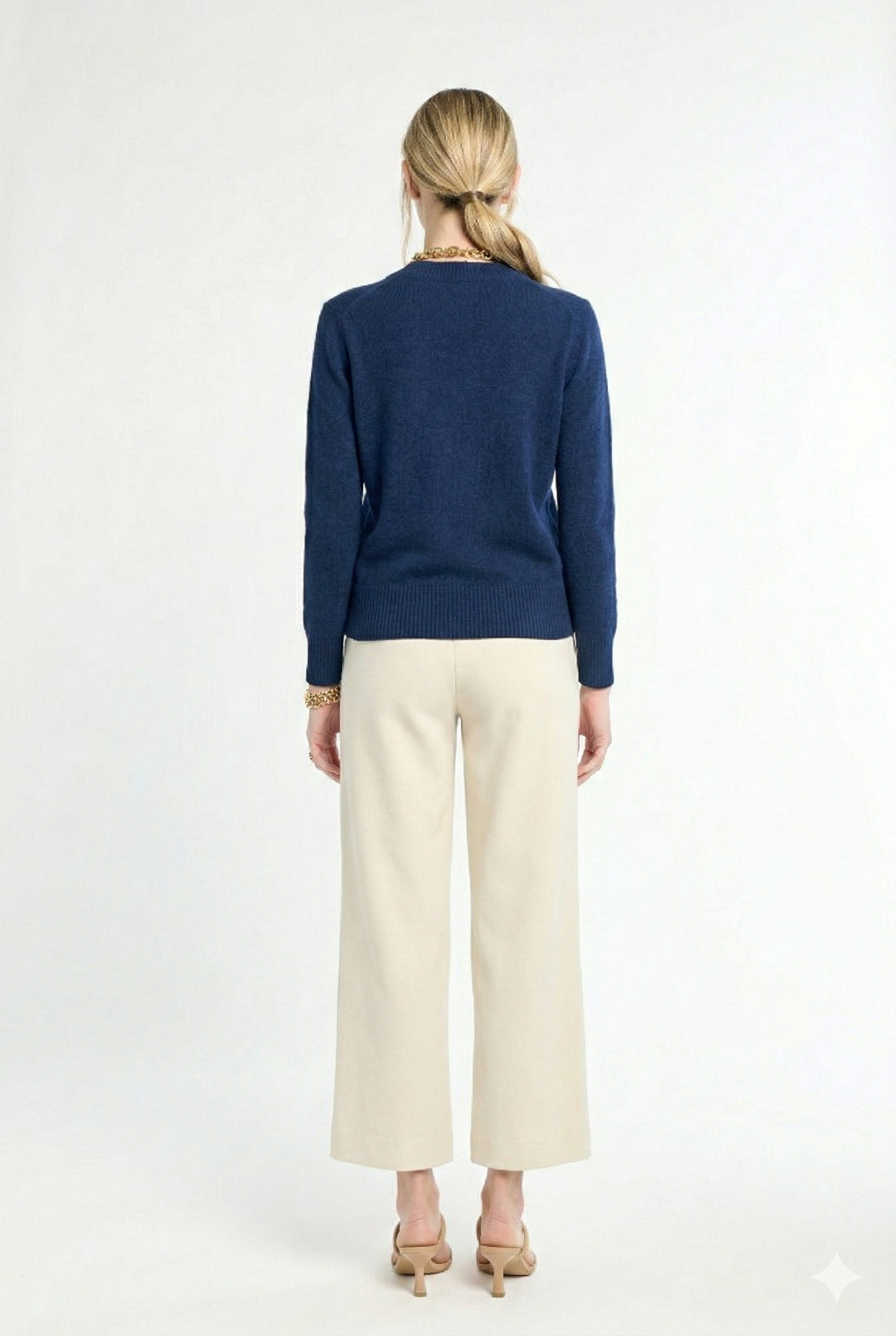 back of woman wearing navy v-neck sweater and cream pants on a white background