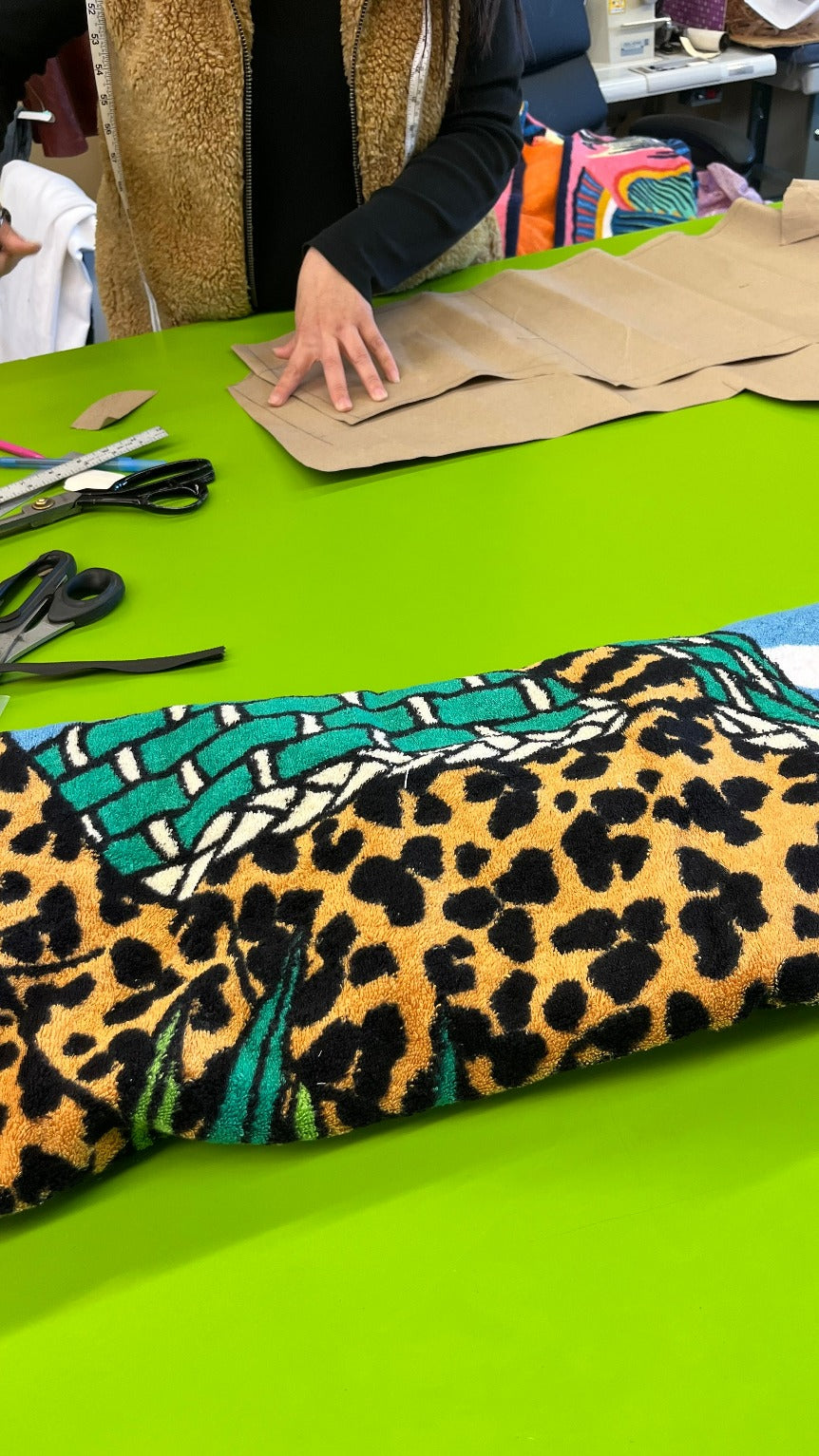 Designer working on fabric with vibrant leopard print and green abstract pattern, alongside scissors, measuring tape, and brown paper patterns.