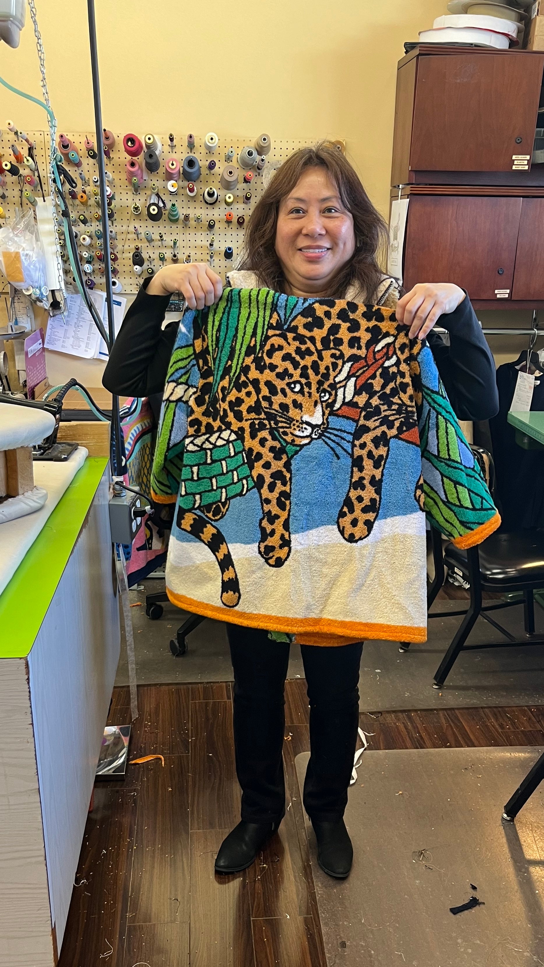 Designer smiling and holding up a towel jacket featuring a leopard print and tropical design, standing in a workspace with sewing supplies