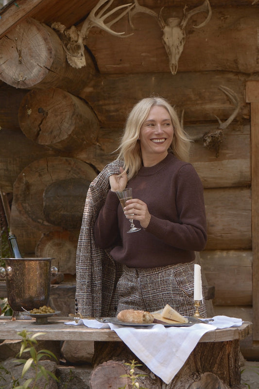 Model wearing the Brown Classic Cashmere Crew paired with plaid trousers, holding a glass of wine in a rustic outdoor setting, near a log cabin.