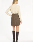 A woman modeling the Tobacco Tweed Meg Mini Skirt with a cream sweater and knee-high boots, back view, photographed against a plain white studio background.