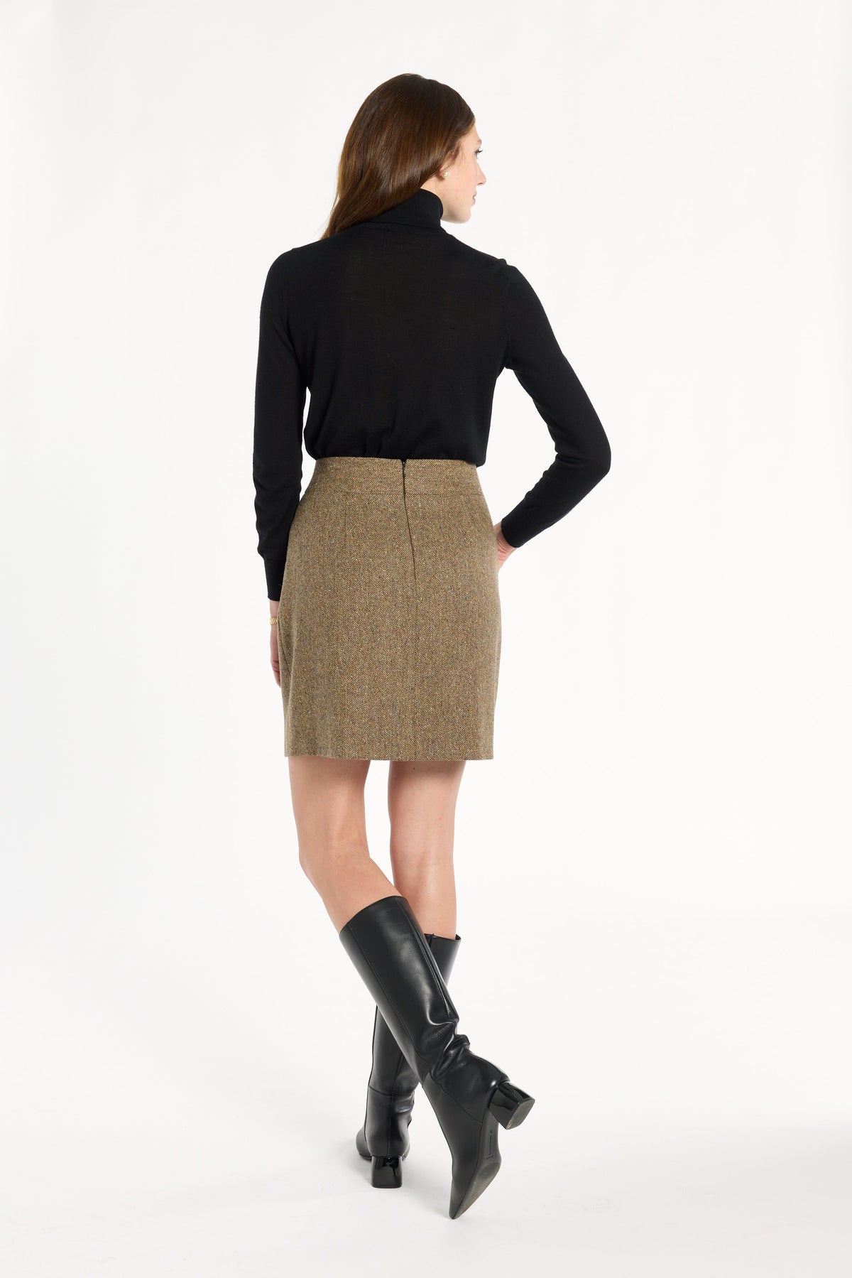 A woman modeling the Painter's Fleck Herringbone Meg Mini Skirt with a black turtleneck and knee-high boots, back view, photographed against a plain white studio background.