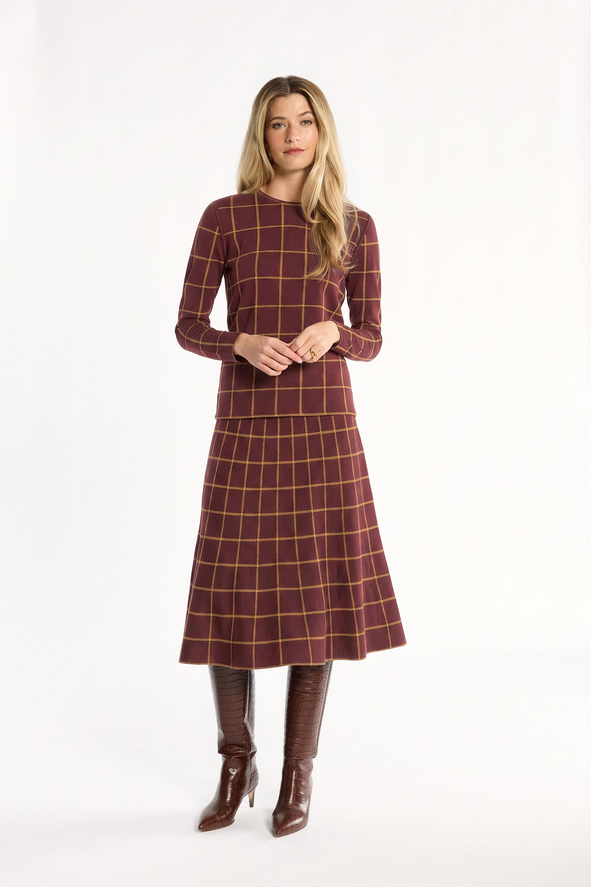 A woman modeling the cranberry side of the Windowpane Plaid Reversible Crewneck with the matching flared skirt and brown boots, front view, photographed against a plain white studio background.
