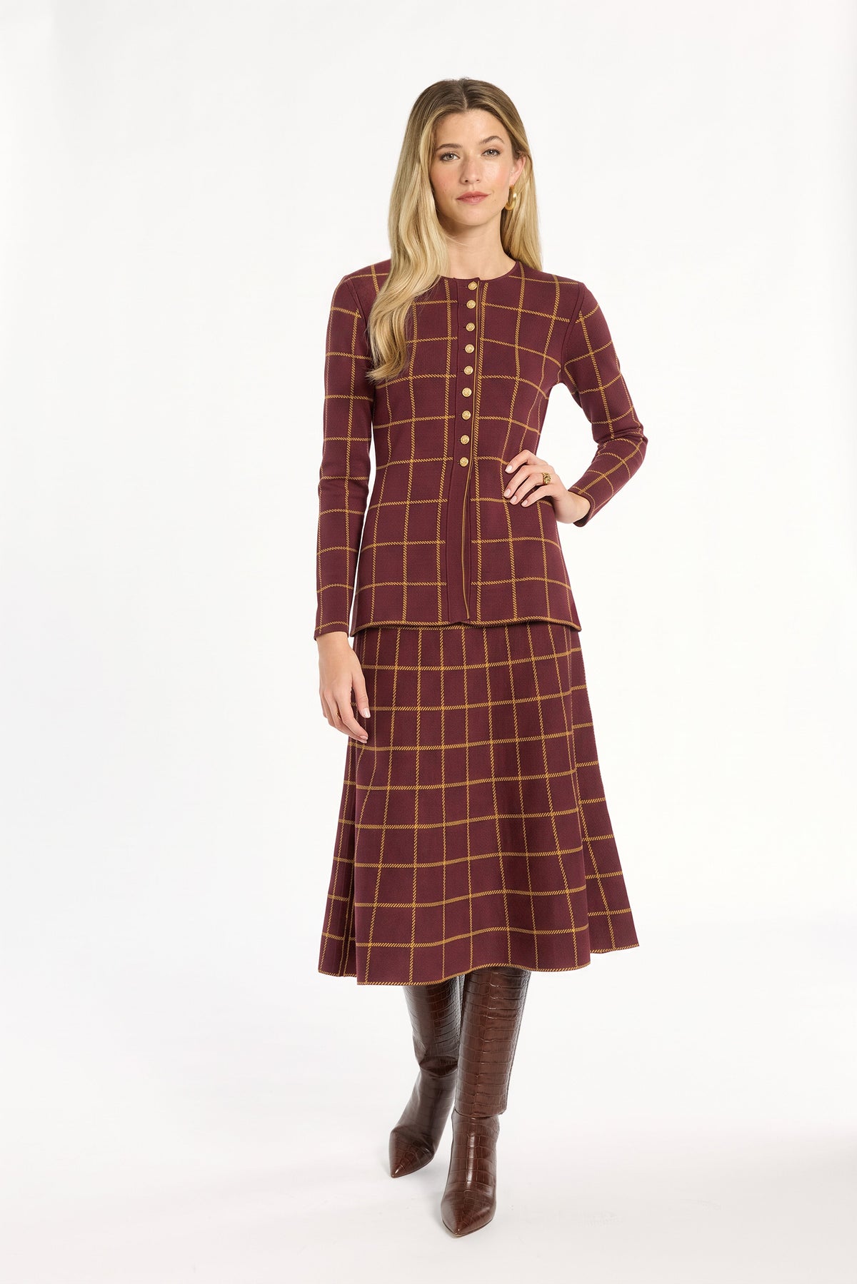 A woman modeling the cranberry side of the Windowpane Plaid Reversible Knit Jacket with the matching flared skirt and brown boots, front view, photographed against a plain white studio background.