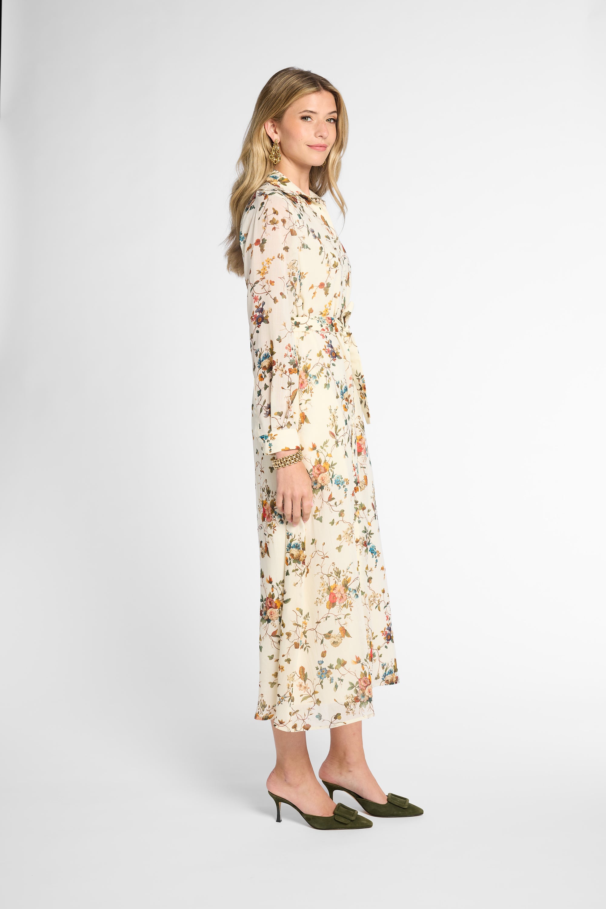A woman modeling a long-sleeved midi dress with a cream base and a dense multicolor floral print, paired with dark green pointed-toe mule heels against a plain light gray studio background.