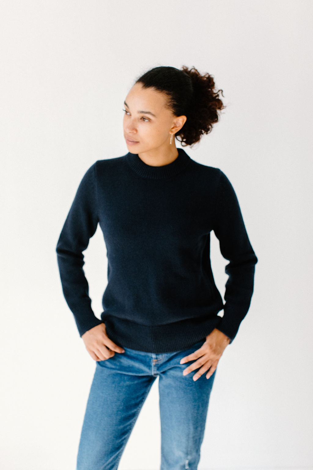 Model wearing the Classic Cashmere Crew in navy, paired with blue jeans, standing in a minimalist setting and looking to the side.