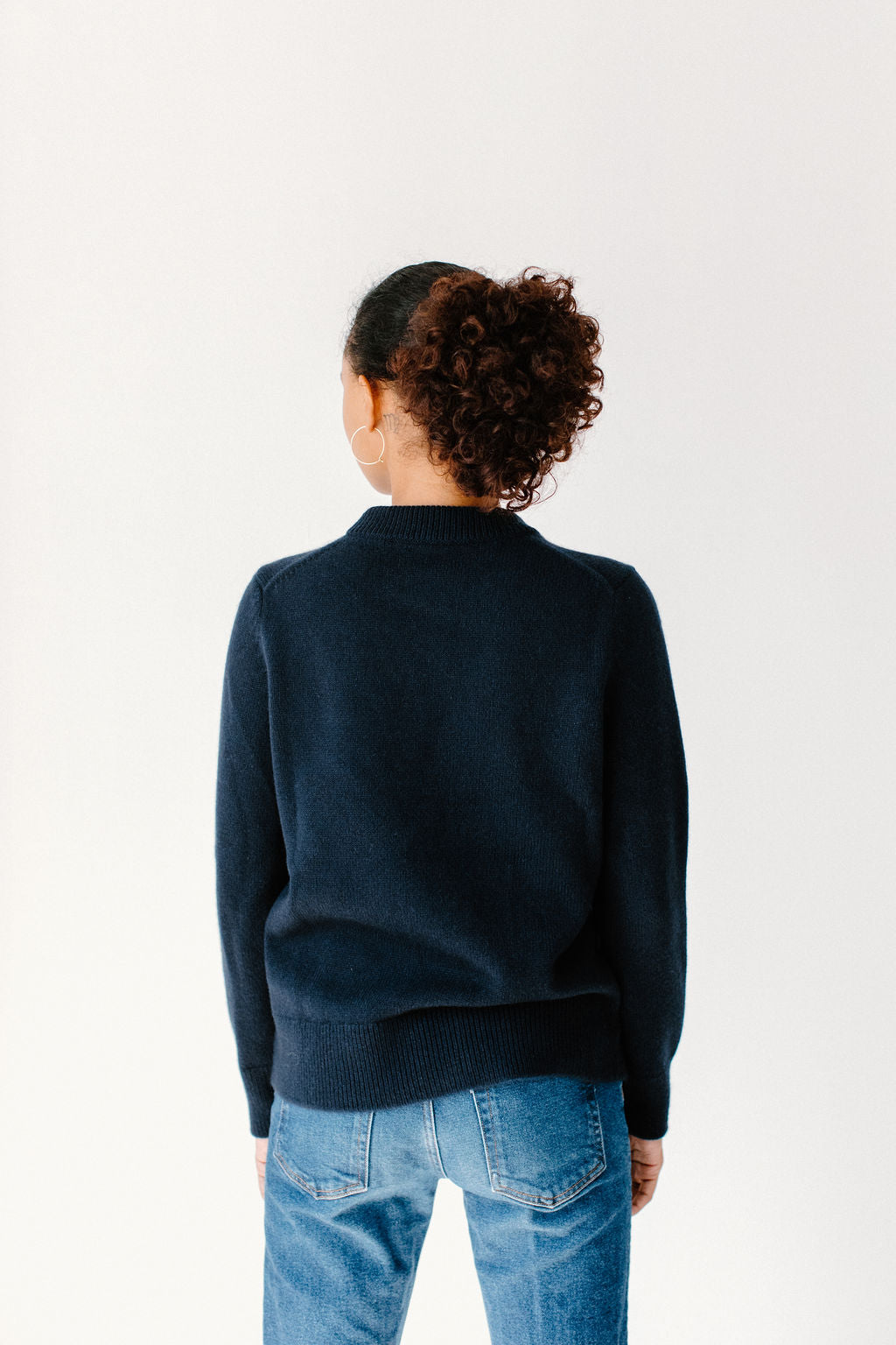 Back view of model wearing the Classic Cashmere Crew in navy, paired with blue jeans, silver hoops, and standing in a minimalist setting.