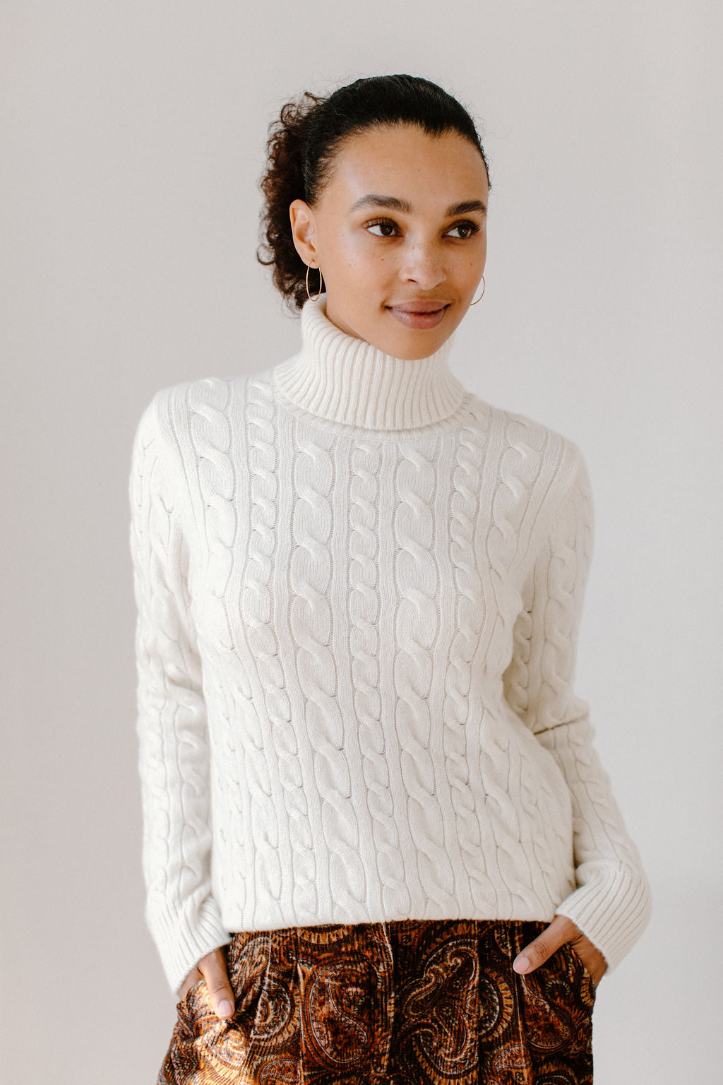 Model wearing the Cream Cable-Knit Turtleneck sweater paired with brown paisley patterned pants, against a white background.