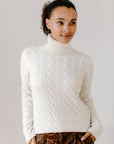Model wearing the Cream Cable-Knit Turtleneck sweater paired with brown paisley patterned pants, against a white background.